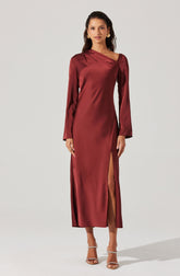 A woman is wearing a long, elegant gown made of shiny satin fabric in a deep burgundy color. The dress features long sleeves, an asymmetrical neckline, and a high slit on one side that reveals her leg. She is also wearing strappy high-heeled sandals.